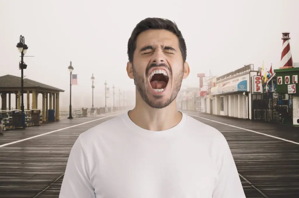 Un homme en chemise blanche se tient sur une promenade vide avec des boutiques et des lampadaires, la bouche grande ouverte comme s'il criait ou hurlait. Le ciel est couvert et l'arrière-plan est brumeux.