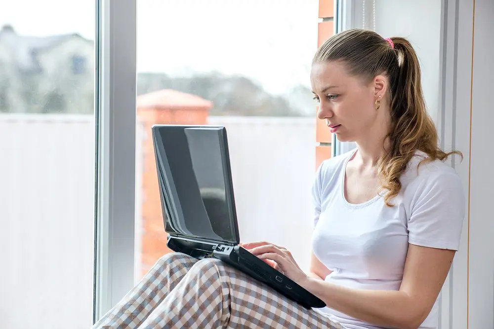 Femme en télétravail sur ordinateur portable près d'une fenêtre.