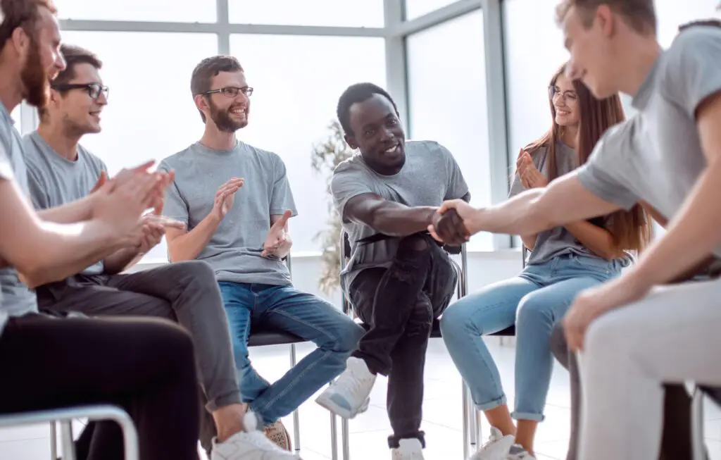 Un groupe de personnes assises en cercle, souriantes, applaudissant, et deux hommes au centre qui tendent la main pour une poignée de main, dans une salle moderne et lumineuse avec de grandes fenêtres.