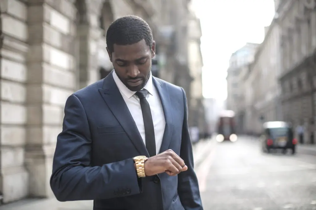 Un homme en costume-cravate bleu marine se tient dans une rue de la ville, regardant sa montre-bracelet en or, avec des bâtiments flous et la circulation en arrière-plan.