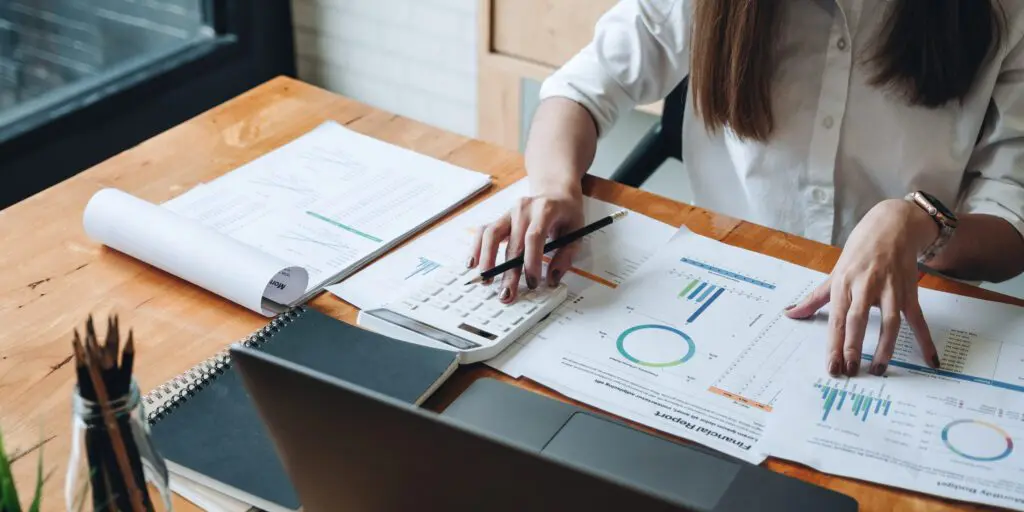 Une personne en chemise blanche est assise à un bureau où se trouvent des tableaux financiers, des graphiques et des documents. Elle utilise une calculatrice et un ordinateur portable, semblant analyser des données ou travailler sur des tâches comptables.
