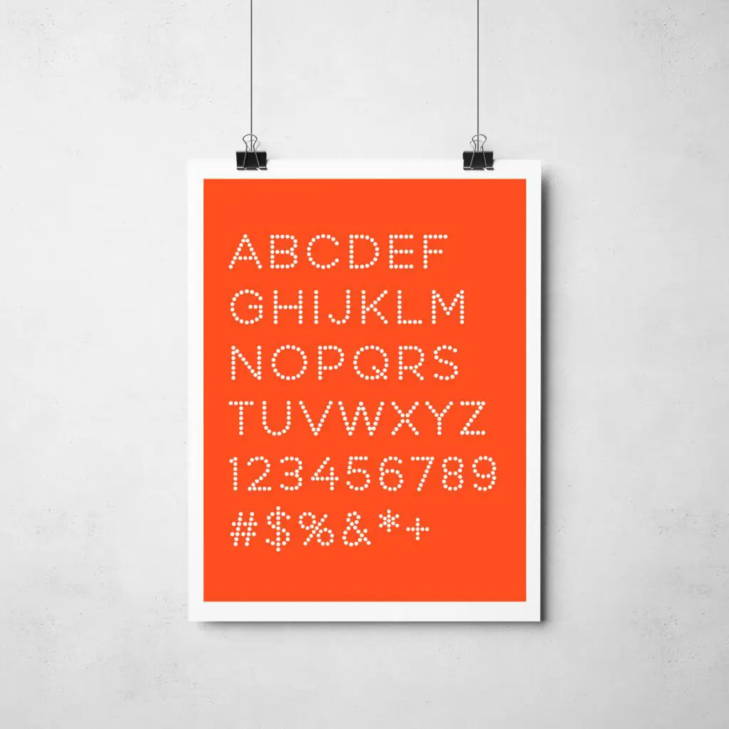 Une affiche avec des lettres majuscules blanches géométriques et pointillées, des chiffres et des symboles sur fond orange est accrochée à un mur clair avec deux pinces noires.