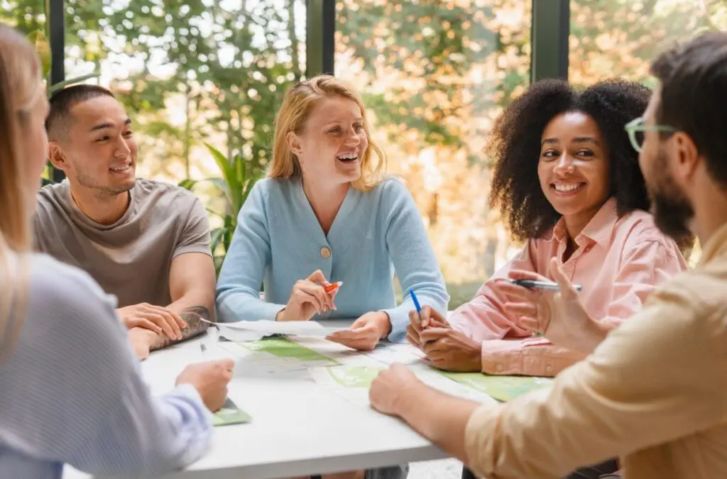 Quatre personnes sont assises autour d'une table avec des papiers et des tableaux, souriant et discutant dans une pièce lumineuse avec de grandes fenêtres et de la verdure à l'extérieur. Elles semblent être en train de discuter ou de se réunir dans un esprit de collaboration.