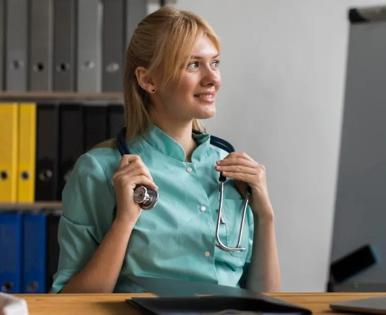 Une jeune femme en blouse médicale est assise à un bureau, souriante et tenant un stéthoscope autour du cou. Des étagères contenant des dossiers colorés et un tableau à feuilles mobiles se trouvent à l'arrière-plan.