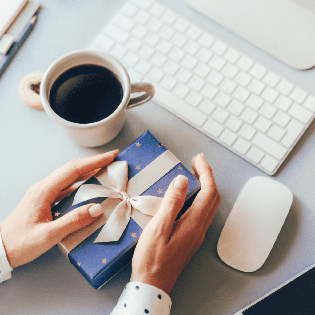 Personne ouvrant un cadeau bleu sur un bureau avec un ordinateur et une tasse de café