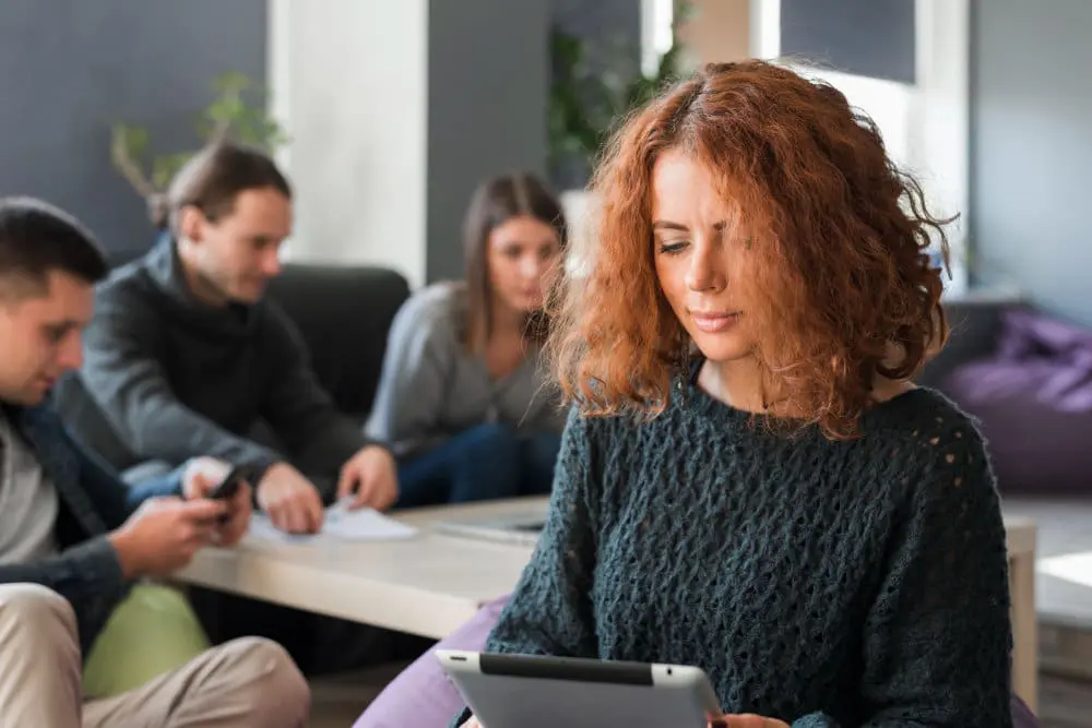 Femme rousse concentrée sur tablette, groupe de collègues en arrière-plan, risques psychosociaux